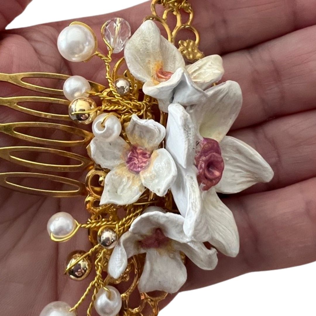 Hand holding candlelight white bridal hair comb with sculpted clay flowers, ivory pearls, and gold vine for scale
