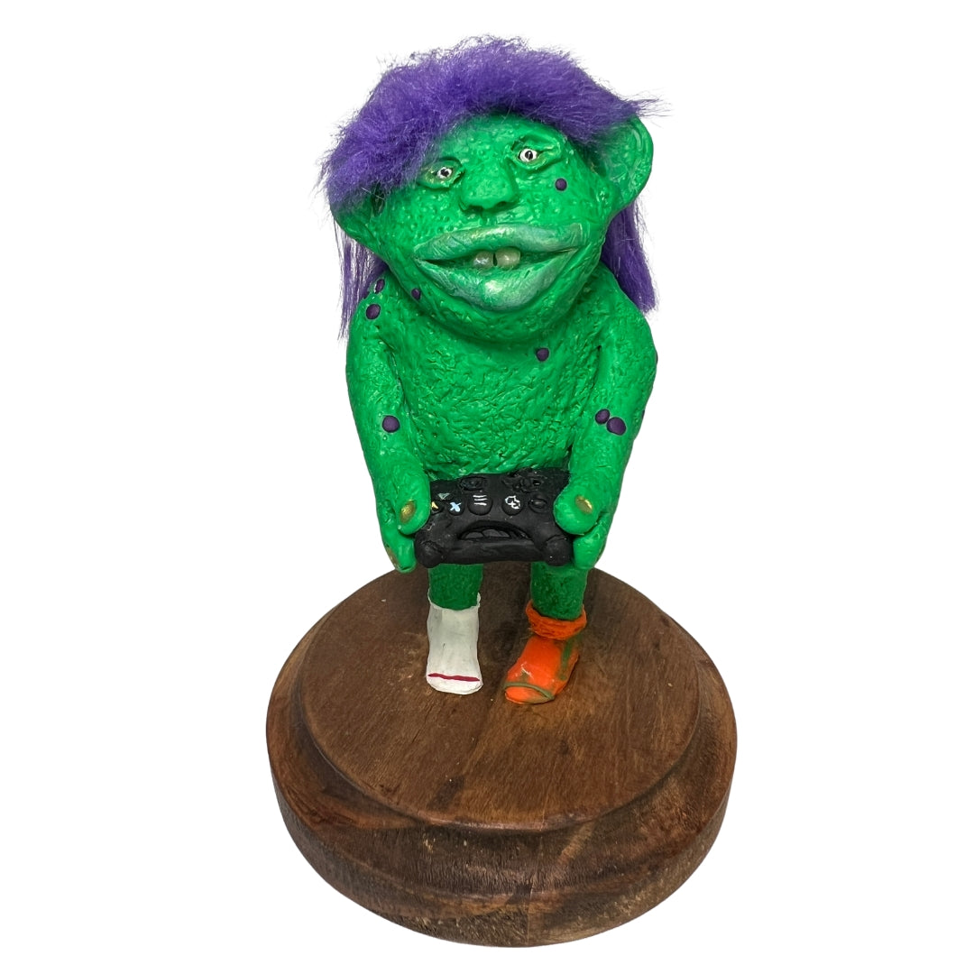 Green Game On gamer creature sculpture with purple hair and a controller on a walnut-stained wooden base.