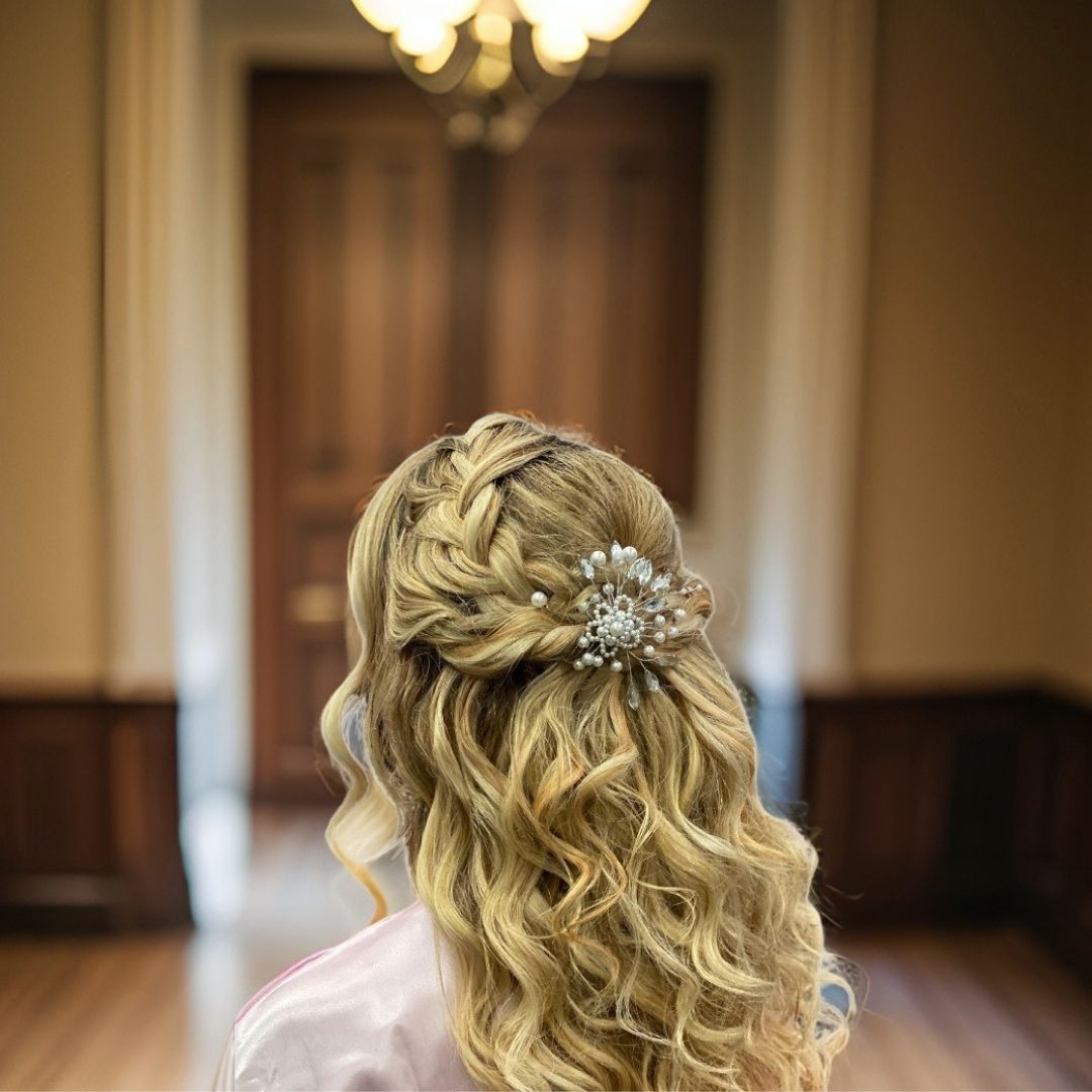 Ivory pearl and crystal bridal hair pin in a braided half up blonde hairstyle with soft curls and chandelier background
