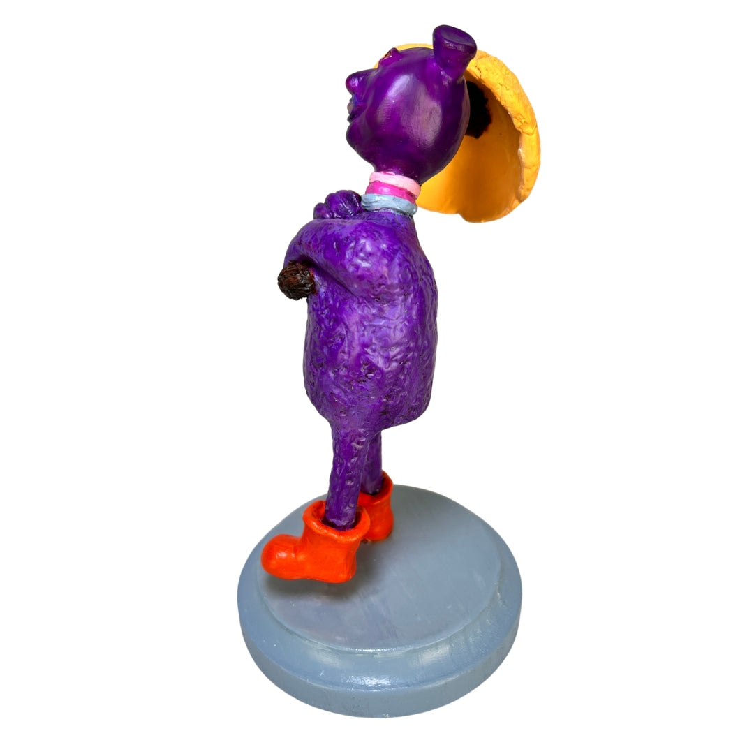 Purple Rainy Day Joy creature sculpture holding a yellow umbrella and wearing orange rain boots on a gray wooden base.