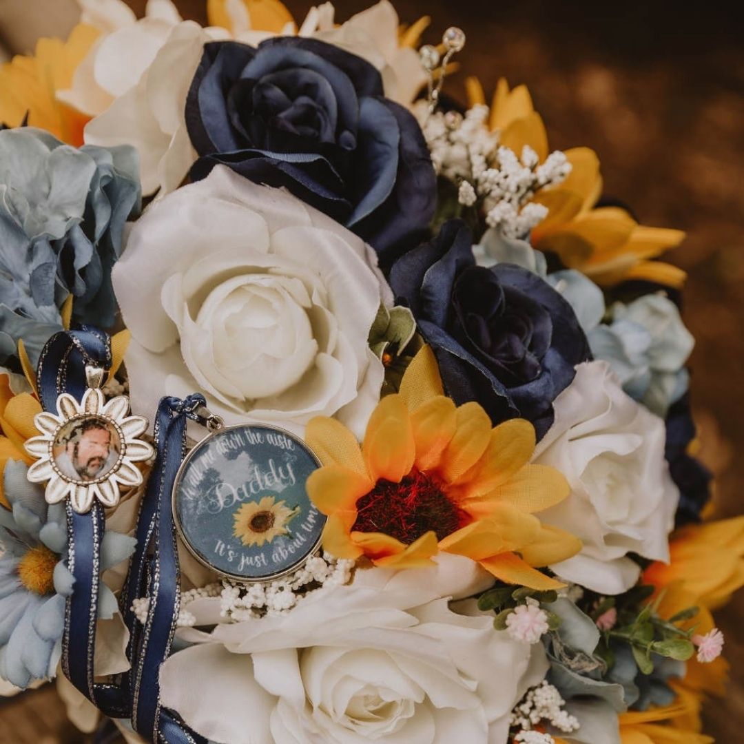 Close-up of cascading bridal bouquet with sunflowers, sapphire blue roses, and ivory roses featuring a memorial photo charm attached with ribbon