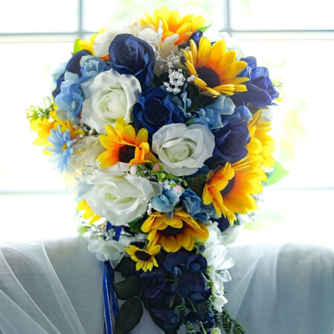Extra large cascading bridal bouquet with sunflowers, sapphire blue roses, ivory roses, and blue hydrangea photographed in natural window light with trailing greenery and optional memorial photo charm