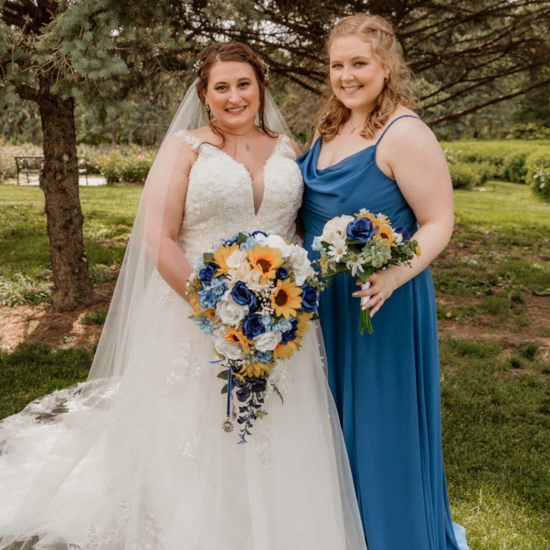 Bride and bridesmaid holding sunflower and sapphire blue cascading bridal bouquet and matching bouquet during outdoor wedding ceremony