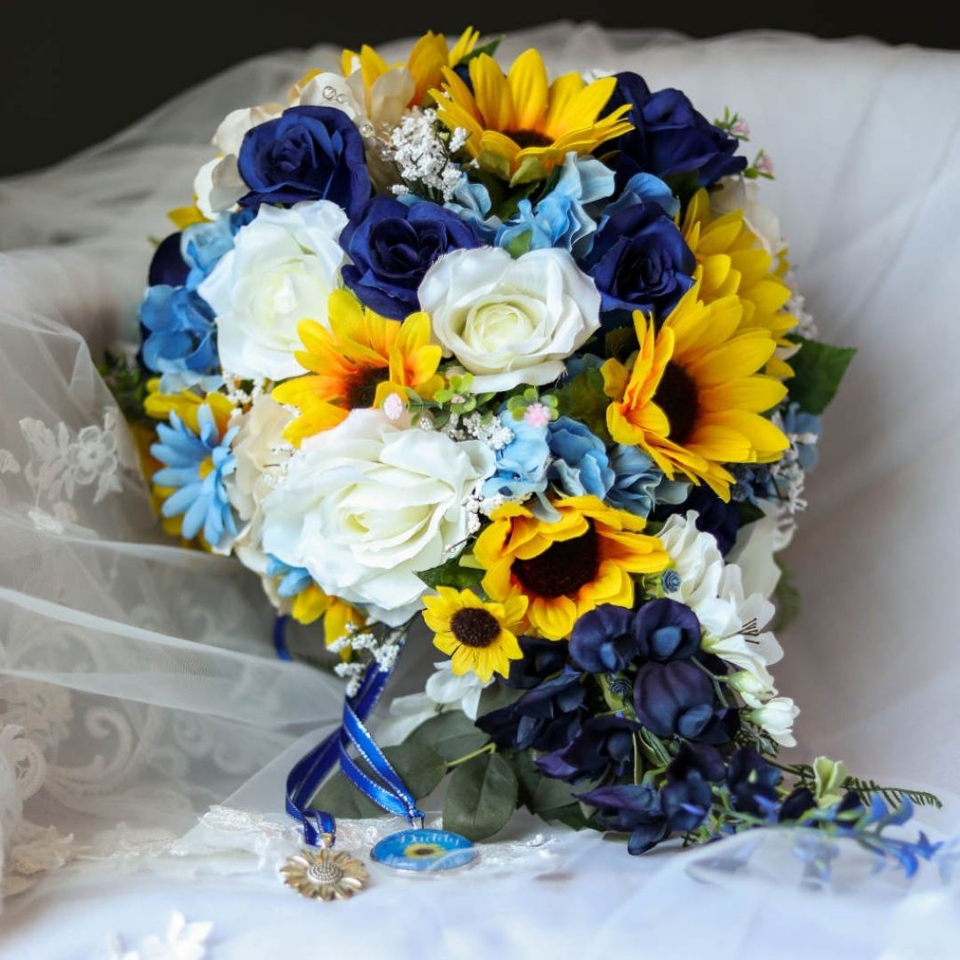 Sunflower and sapphire blue cascading bridal bouquet resting on veil with ivory roses, blue hydrangea, and memorial charm detail