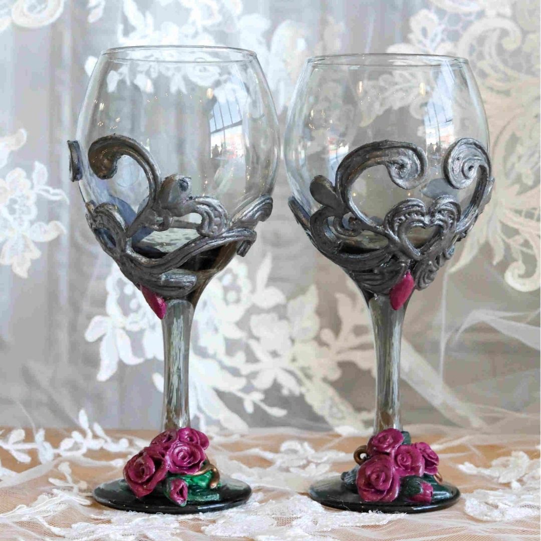 Set of two Victorian-inspired gothic wine glasses with sculpted silver scrollwork and burgundy roses on the bases.