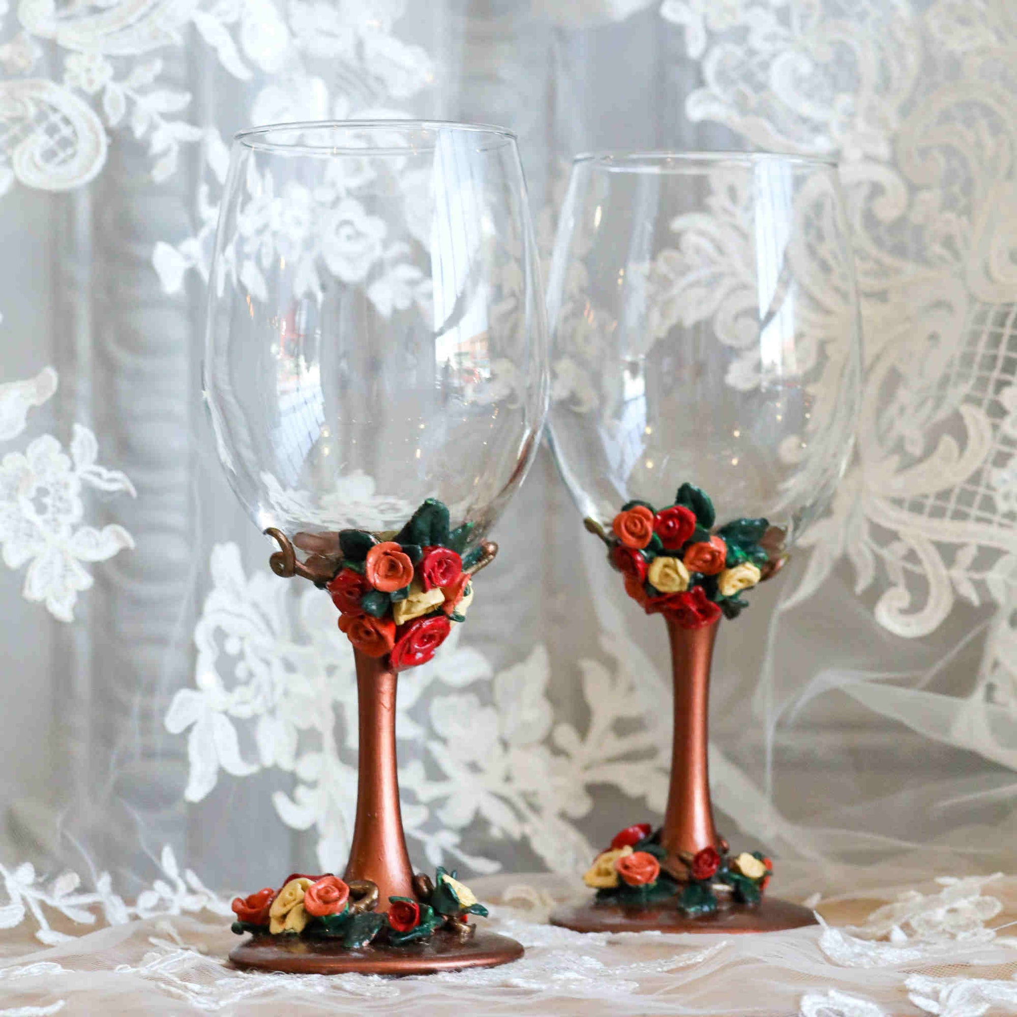 Each wine glass sits atop of rich metallic copper stem and is adorned with polymer clay flowers in red, orange, and yellow flowers within green leaves and vines.