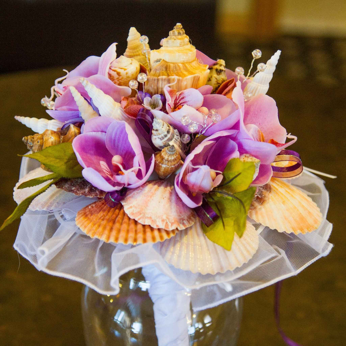 Bring the beauty of the beach to your tropical wedding ceremony. This cascading bouquet is adorned with various shells, silk orchids, ribbon, greenery and crystal bead stems.