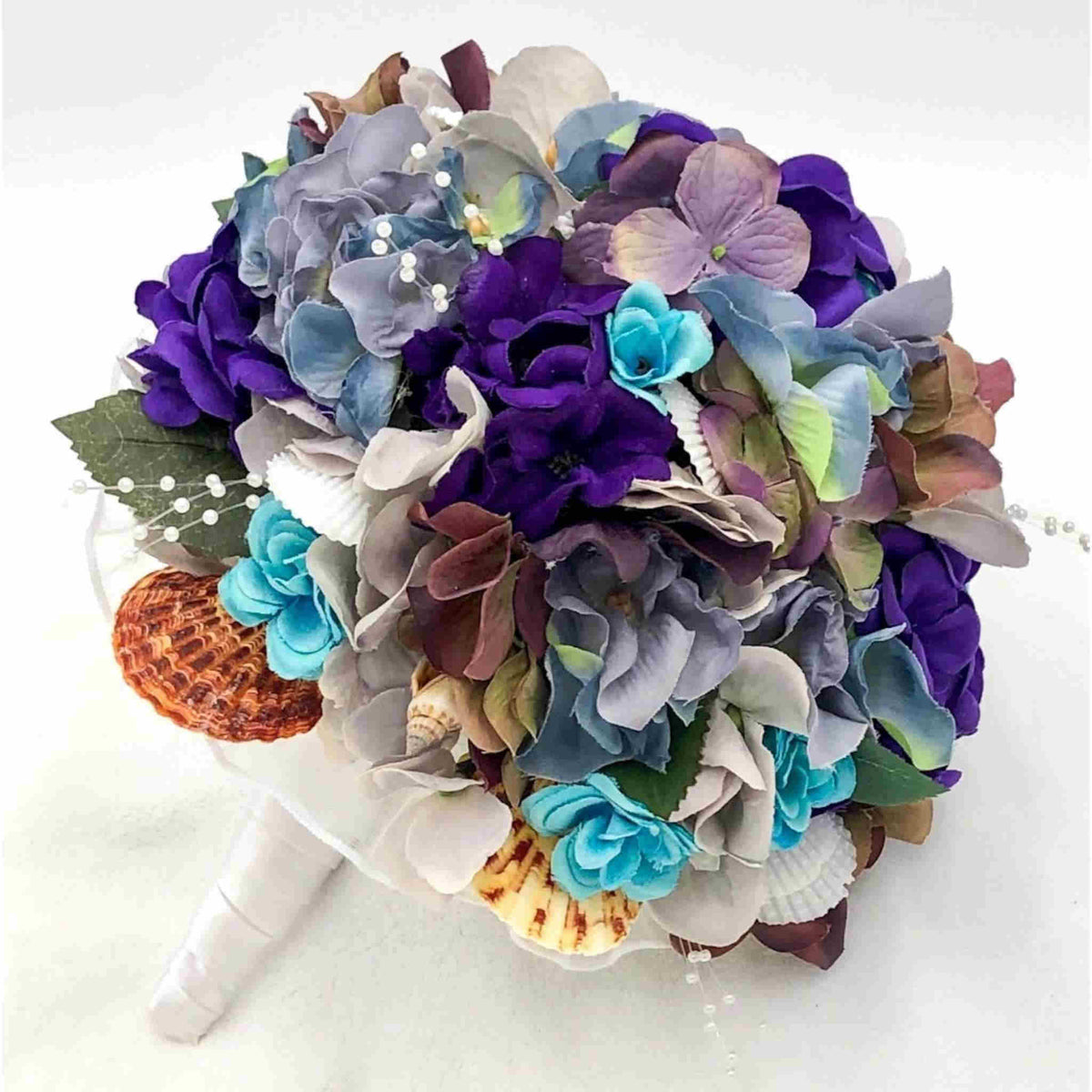 Made with purple and turquoise flowers, accented with seashells and white pearl stems, this beautiful bouquet sits atop a handle with a comfort grip wrapped in white satin with teal accent ribbon.