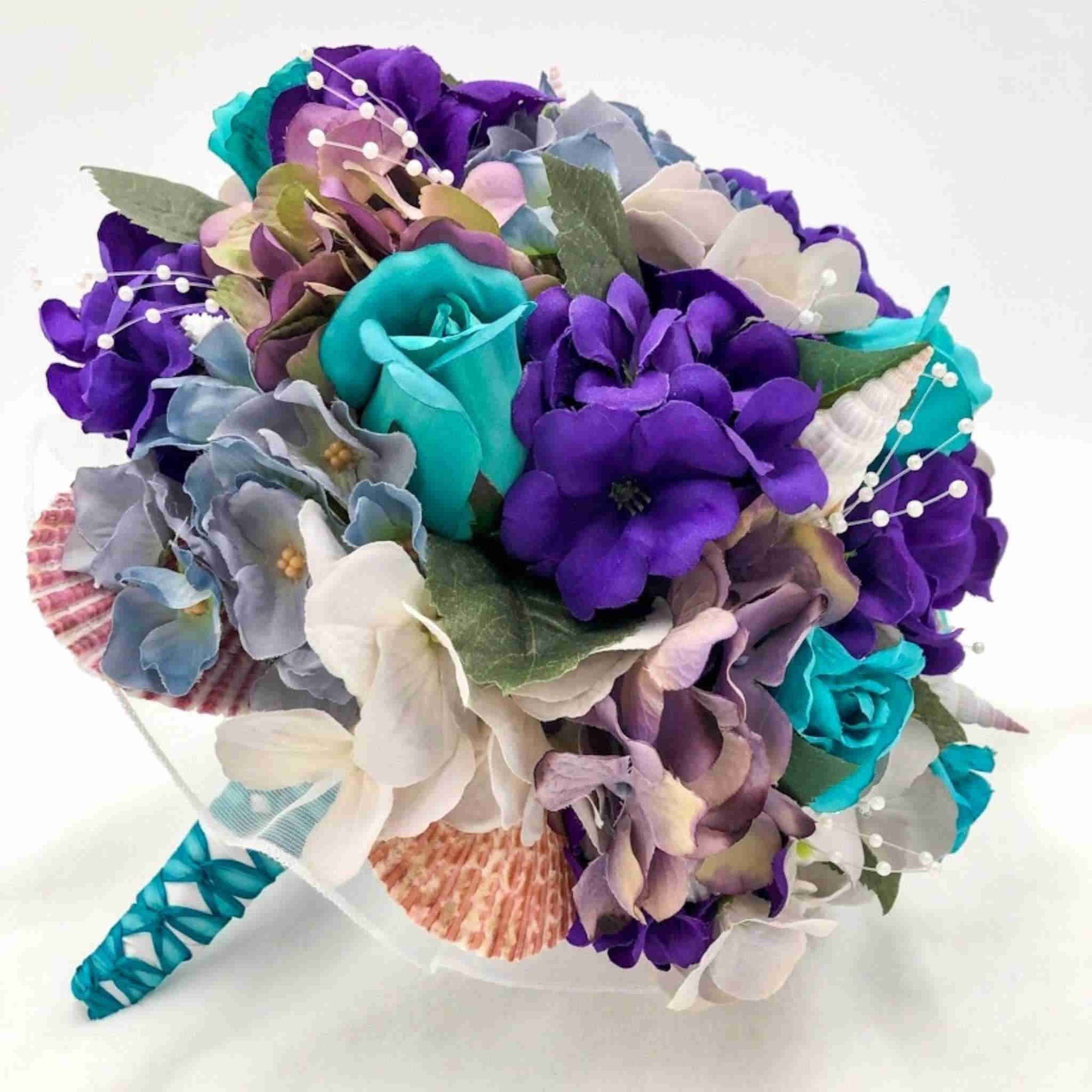 Made with purple and turquoise flowers, accented with seashells and white pearl stems, this beautiful bouquet sits atop a handle with a comfort grip wrapped in white satin with teal accent ribbon.