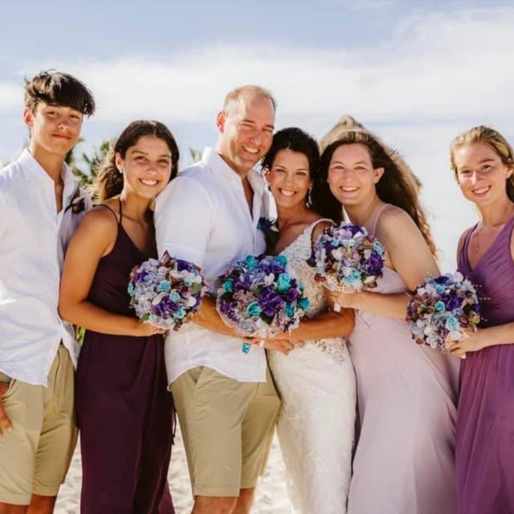 Made with purple and turquoise flowers, accented with seashells and white pearl stems, this beautiful bouquet sits atop a handle with a comfort grip wrapped in white satin with teal accent ribbon.