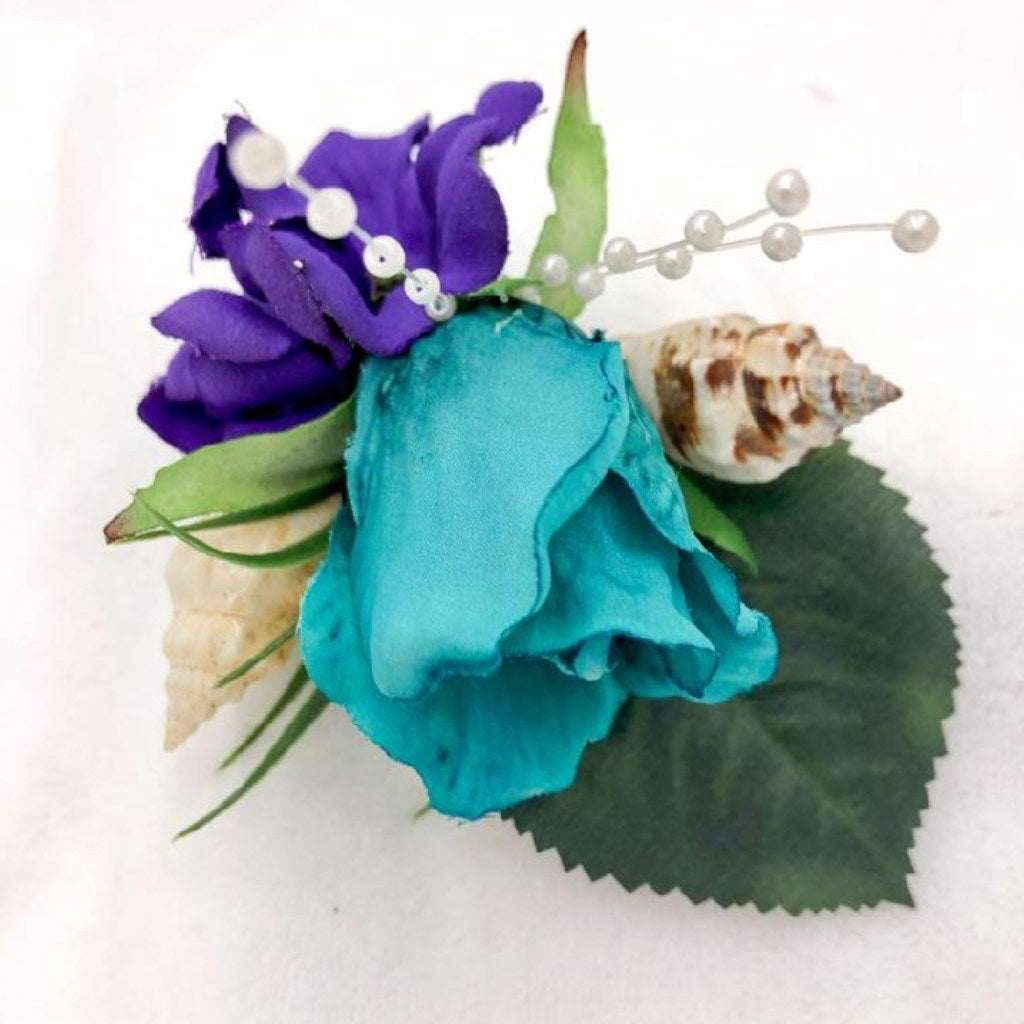 This tropical accessory is made with purple and turquoise flowers, accented with seashells and tiny pearls. The base of this boutonniere is wrapped in ribbon for an extra pop of color.