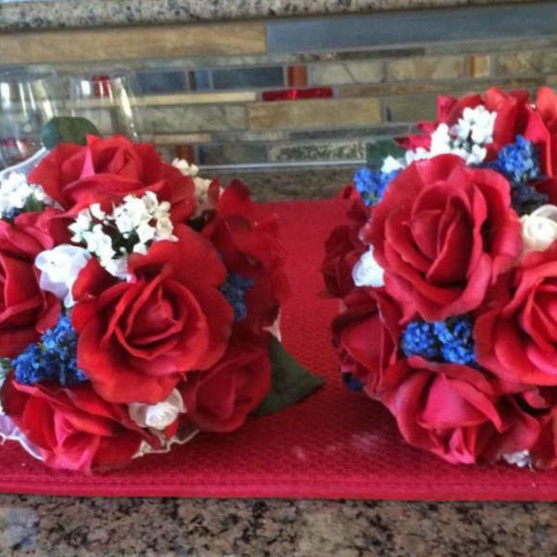 This round bouquet is lush with red silk flowers, accented with small blue and white flowers, and sits atop a plastic handle, wrapped in ribbon, finished with a simple lace collar.