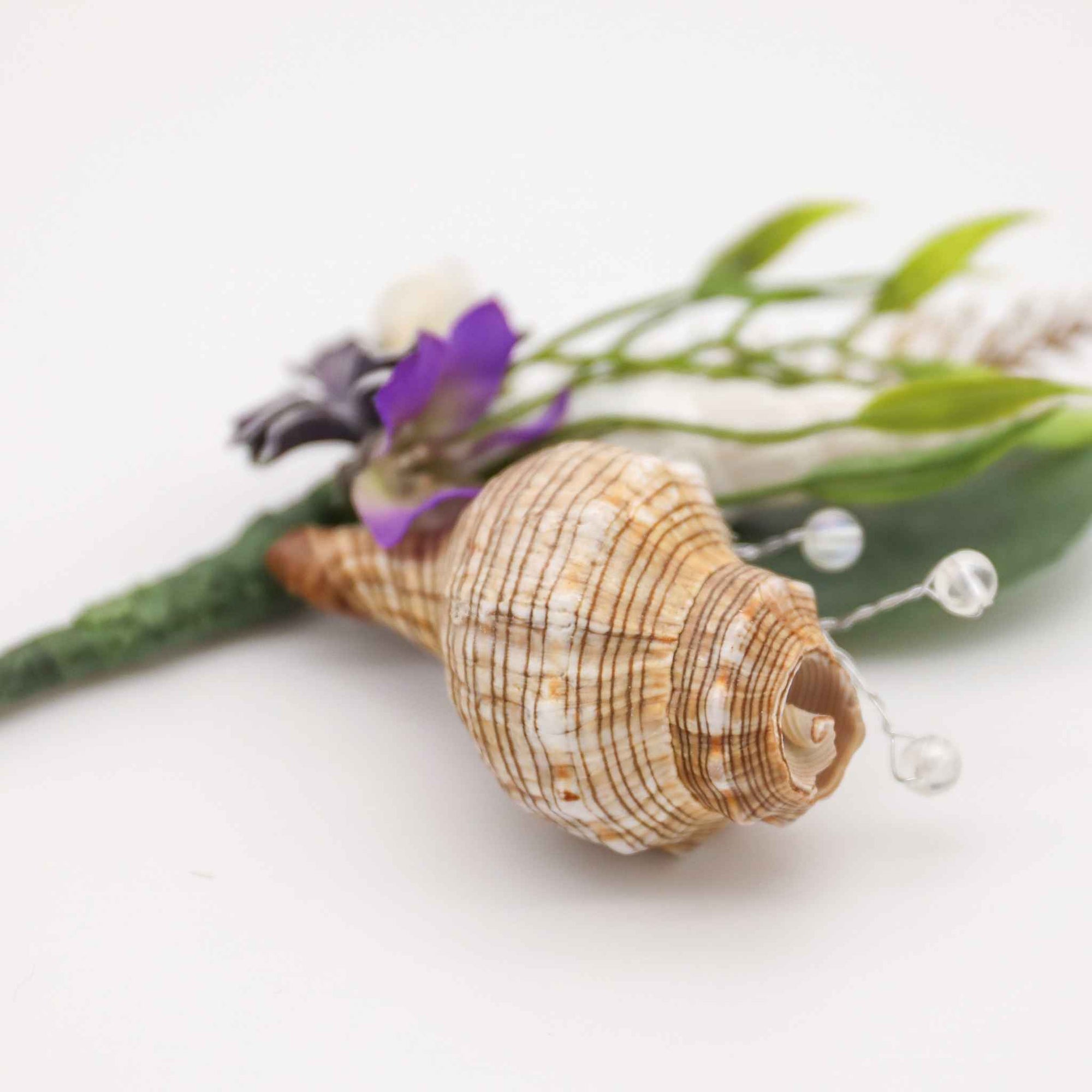 Bring the beach to your summer or tropical wedding ceremony. Set in greenery and adorned with cone shells and some small silk flowers, this boutonniere is a great accessory for any summer, beach, or tropical wedding.