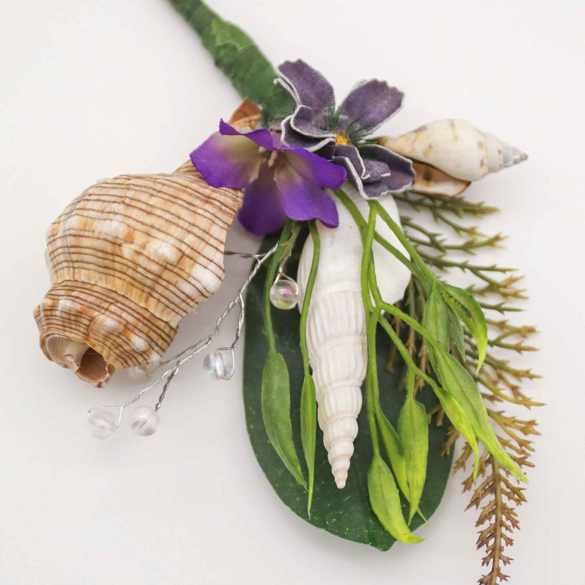 Bring the beach to your summer or tropical wedding ceremony. Set in greenery and adorned with cone shells and some small silk flowers, this boutonniere is a great accessory for any summer, beach, or tropical wedding.